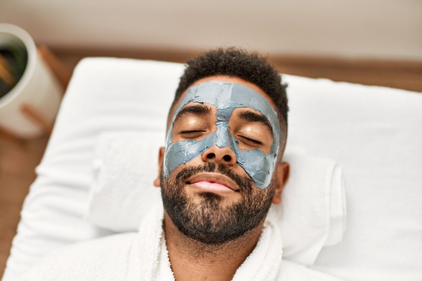 A man lays face up on a spa table, his face covered with a grey cream.