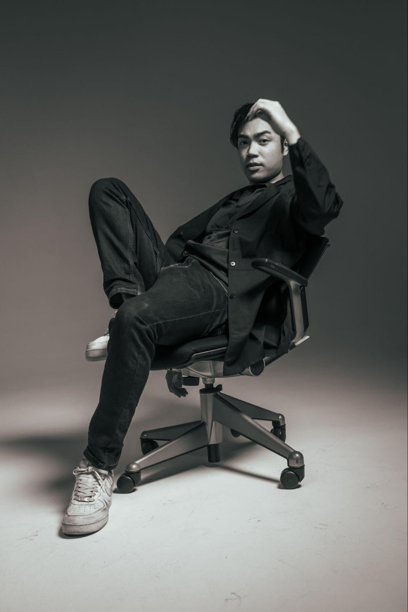 Black and white photo of a person leaning back on a rolling office chair as they look into the camera.