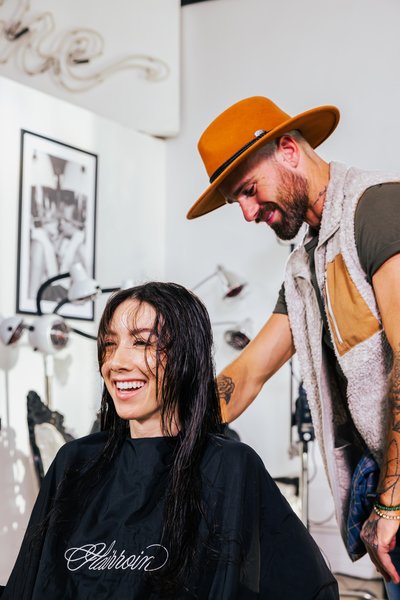 A smiling client sits in a salon chair with wet hair while a stylist prepares to cut their hair.