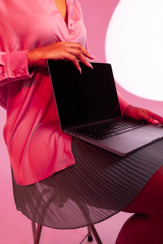 A person wearing a pink shirt and gray skirt sits on a stool, a laptop with a black screen on their lap.