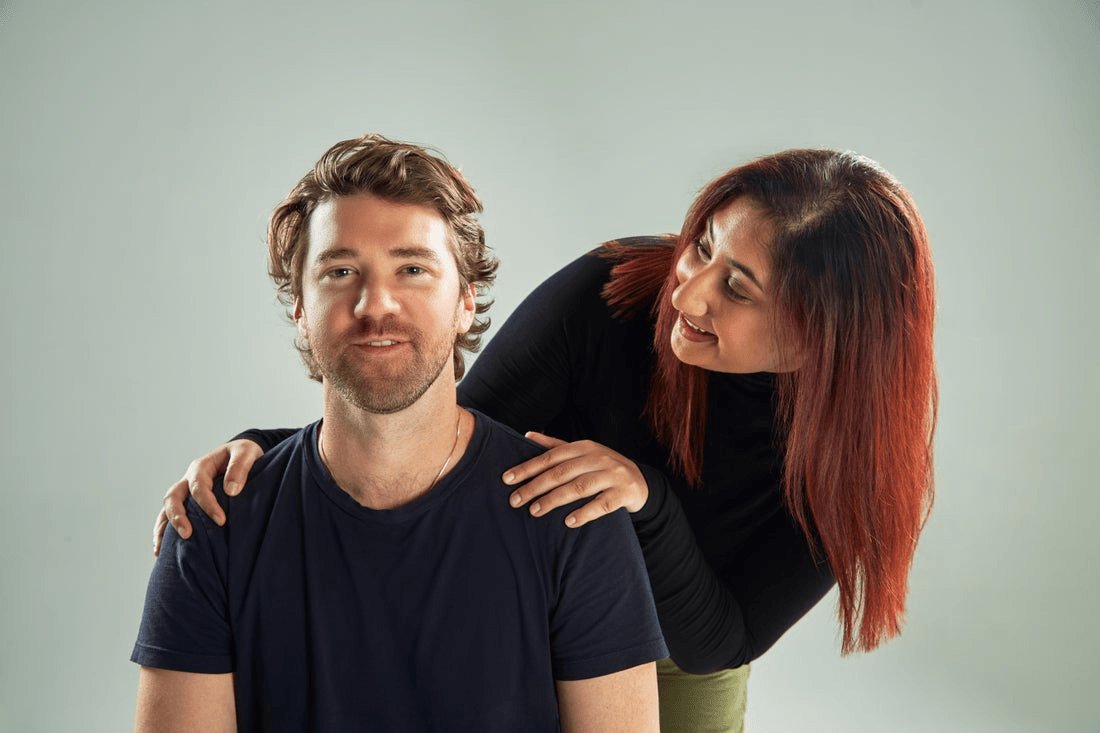 A sitting man looks forward while a woman, standing slightly behind him, smiles at him and holds his shoulders.